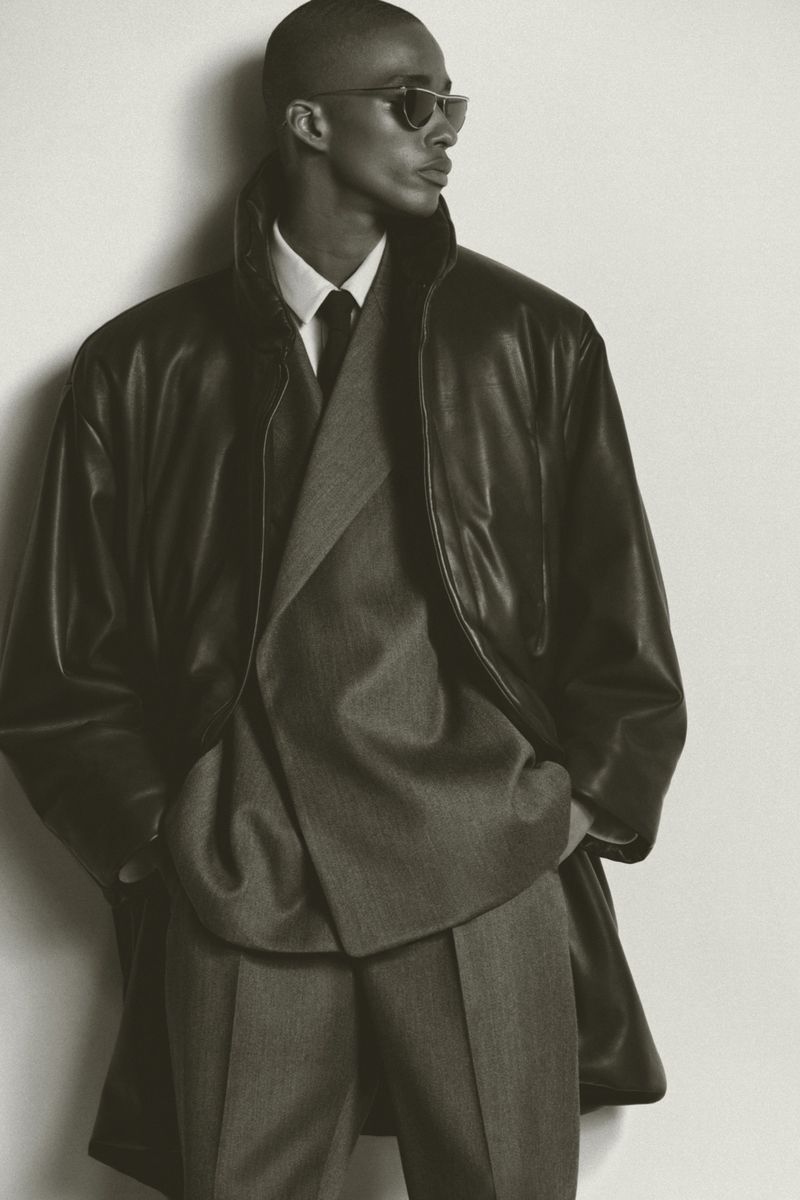 Elegant man in a grey suit and black leather coat posing in front of a white wall, showcasing classic and sophisticated menswear.