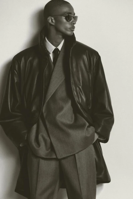 Elegant man in a grey suit and black leather coat posing in front of a white wall, showcasing classic and sophisticated menswear.