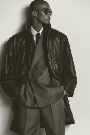 Elegant man in a grey suit and black leather coat posing in front of a white wall, showcasing classic and sophisticated menswear.