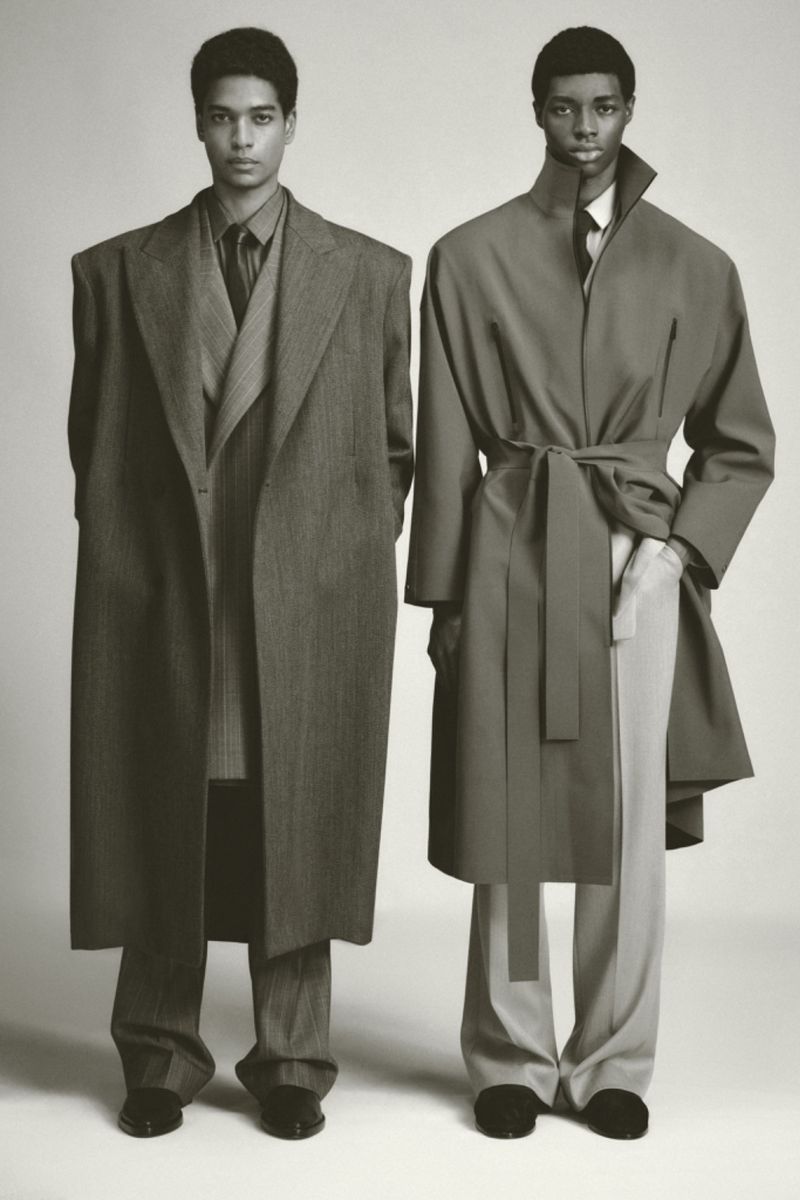 Two adult men in overcoats and trousers stand in front of a neutral background, showcasing fashion with a muted color palette and soft lighting.