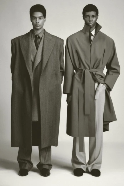 Two adult men in overcoats and trousers stand in front of a neutral background, showcasing fashion with a muted color palette and soft lighting.