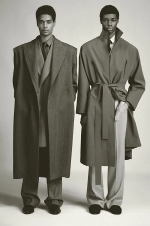 Two adult men in overcoats and trousers stand in front of a neutral background, showcasing fashion with a muted color palette and soft lighting.