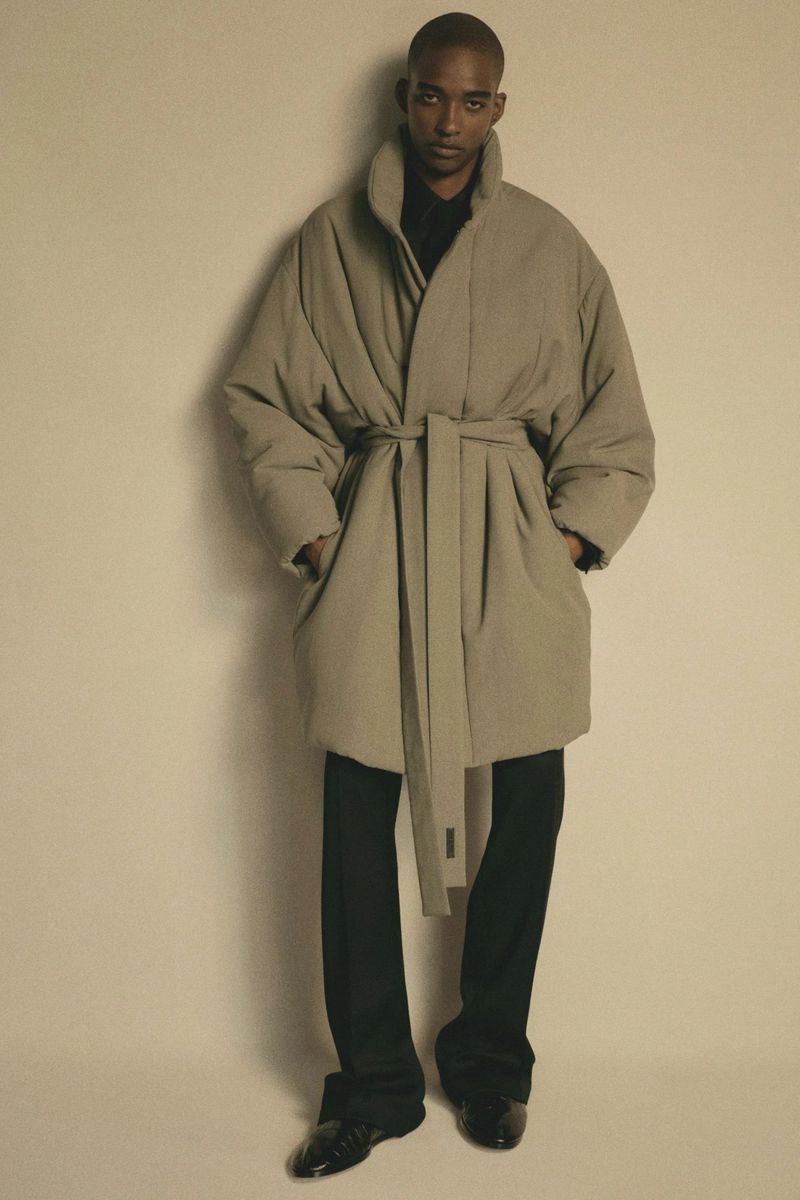 Adult male with dark skin poses in a beige belted puffer coat and black trousers against a beige background in a studio setting.