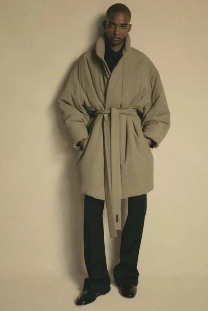 Adult male with dark skin poses in a beige belted puffer coat and black trousers against a beige background in a studio setting.