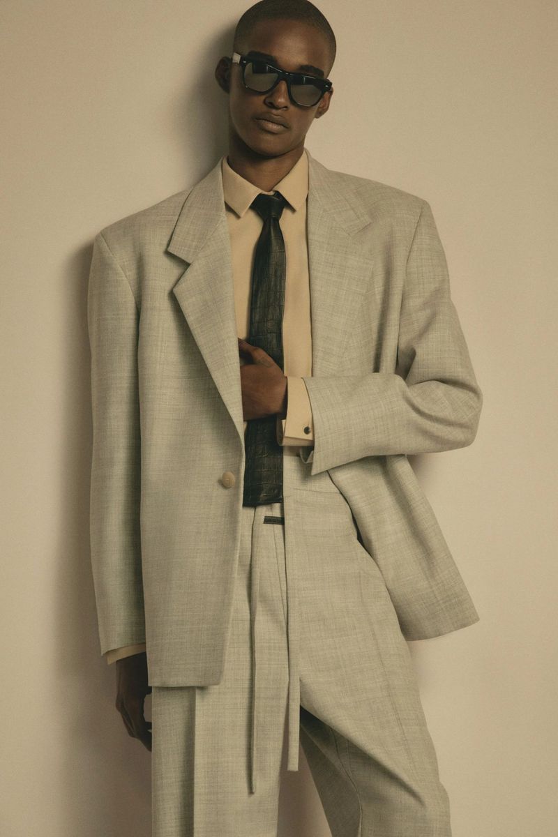 Model in grey tailored suit jacket and matching trousers with dark sunglasses and black tie against a seamless backdrop