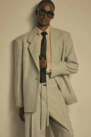 Model in grey tailored suit jacket and matching trousers with dark sunglasses and black tie against a seamless backdrop