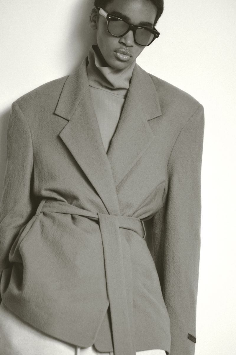 Studio portrait of a woman with sunglasses wearing a light-gray blazer and turtle neck sweater against a white background.