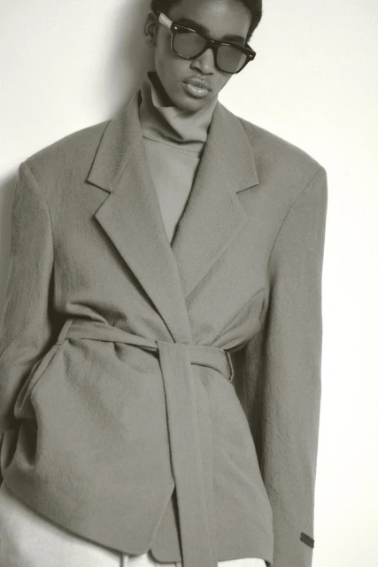 Studio portrait of a woman with sunglasses wearing a light-gray blazer and turtle neck sweater against a white background.
