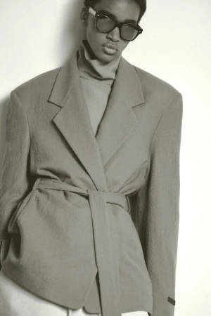 Studio portrait of a woman with sunglasses wearing a light-gray blazer and turtle neck sweater against a white background.