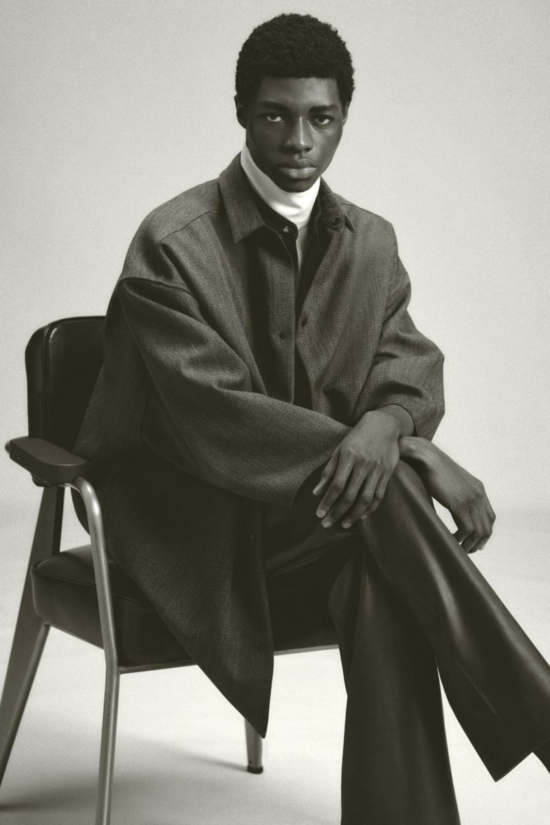 An African-American adult male sits on an arm chair in a studio setting, wearing a gray overcoat and dark pants against a neutral background.