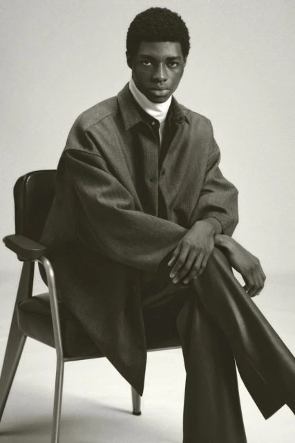 An African-American adult male sits on an arm chair in a studio setting, wearing a gray overcoat and dark pants against a neutral background.