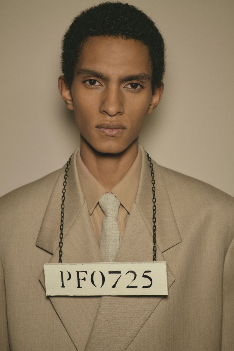 Portrait of a male model wearing a beige suit, matching shirt and tie, and an identification sign hung from his neck in a photo studio.