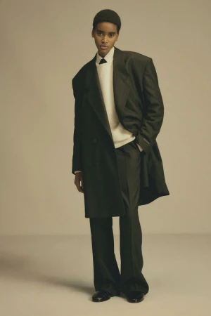 Model wearing a dark oversized coat over a white sweater against a plain neutral background