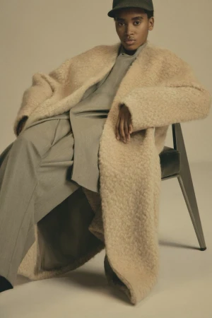 Model wearing a textured beige coat and baseball cap seated on a chair against a neutral background for Fear of God.