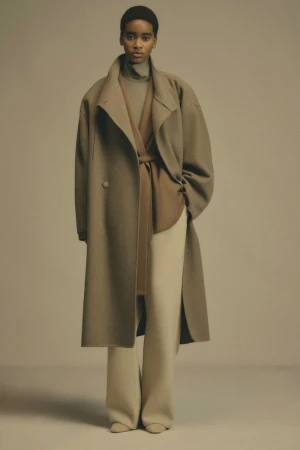 Model wearing an oversized camel wool coat and light trousers standing against a neutral studio backdrop