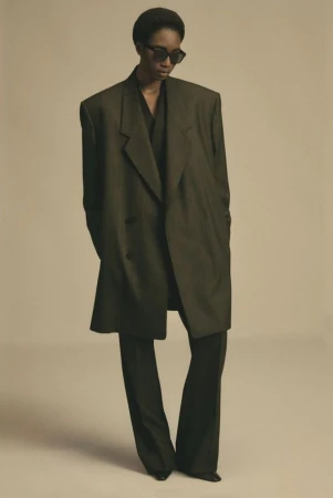 Model wearing an oversized dark blazer and sunglasses standing against a neutral studio background