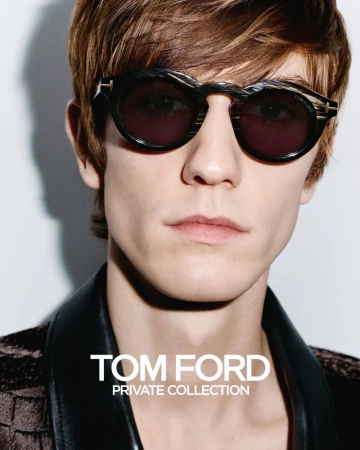 Close-up portrait of a brown-haired male model wearing decorative sunglasses and a dark textured jacket in a fashion setting.