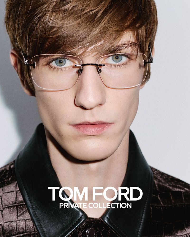 Close-up of a fair-skinned adult male model with light brown hair and blue eyes wearing rimless eyeglasses against a white background.