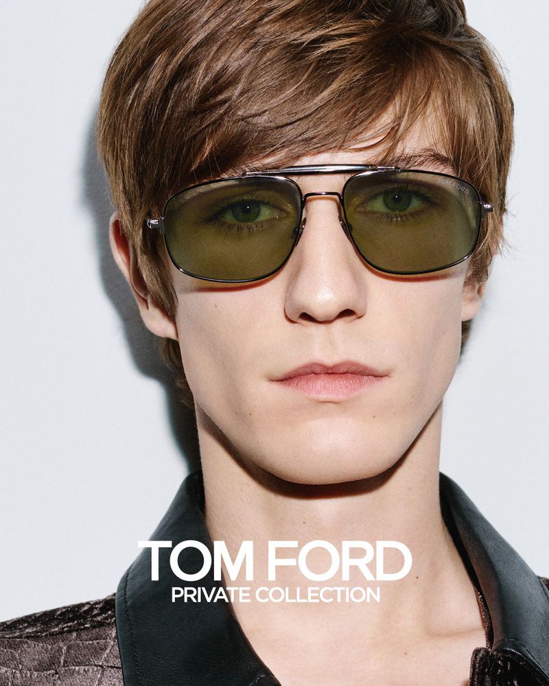 Close-up portrait of a young male model wearing tinted sunglasses and a textured dark jacket against a white wall.
