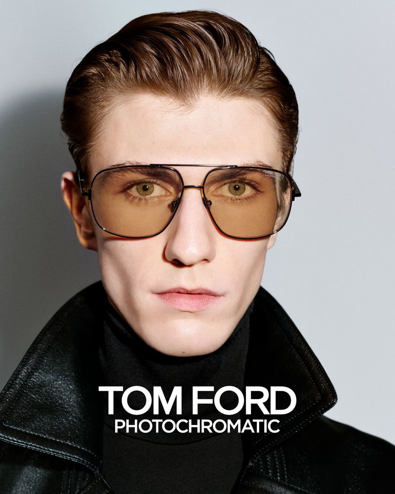 Close-up portrait of a male model wearing tinted aviator sunglasses and a black leather jacket against a grey backdrop.