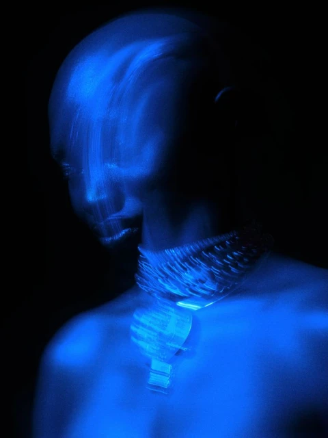 Abstract long exposure of an adult wearing a chunky chainlink necklace with a pendant in electric blue against a dark background.