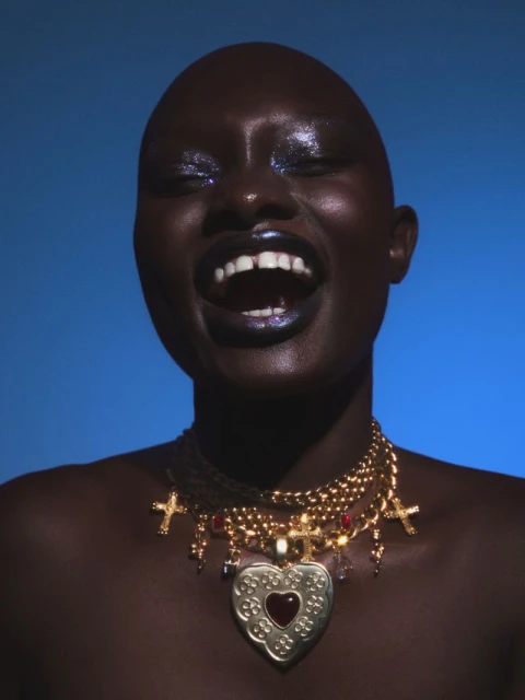 Close-up of Black woman with shaved head smiling against a blue backdrop, adorned with gold chain necklaces and glitter eye makeup.