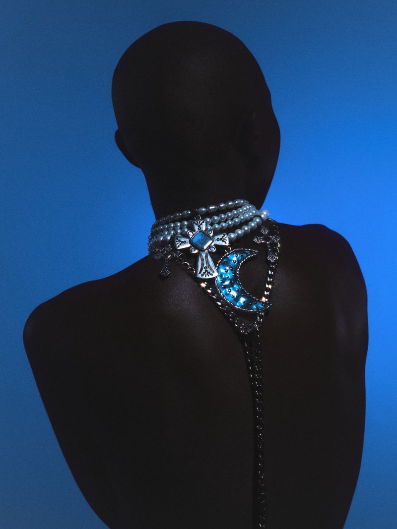 Back view of a person with bare skin, wearing an elaborate pearl and gemstone necklace against a gradient blue background.