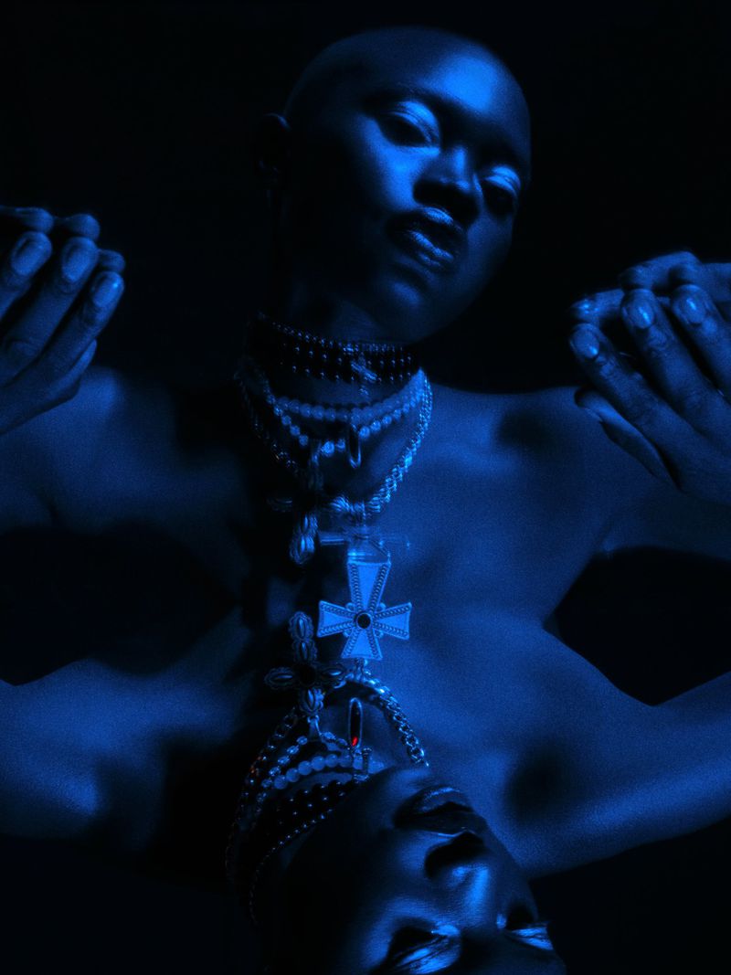 Bald adult Black woman in a blue studio setting, adorned with multiple necklaces, is reflected below. The lighting is dramatic.