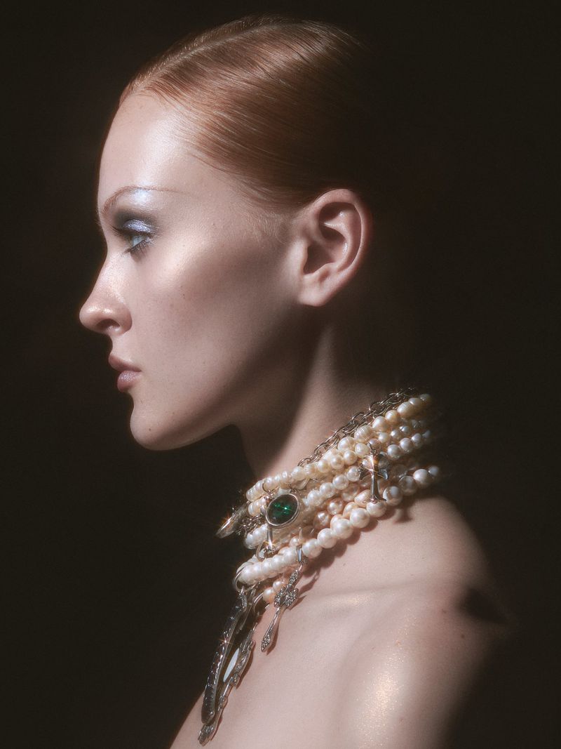 Profile shot of a woman with red hair wearing a pearl and emerald necklace, styled with blue eyeshadow against a dark background.