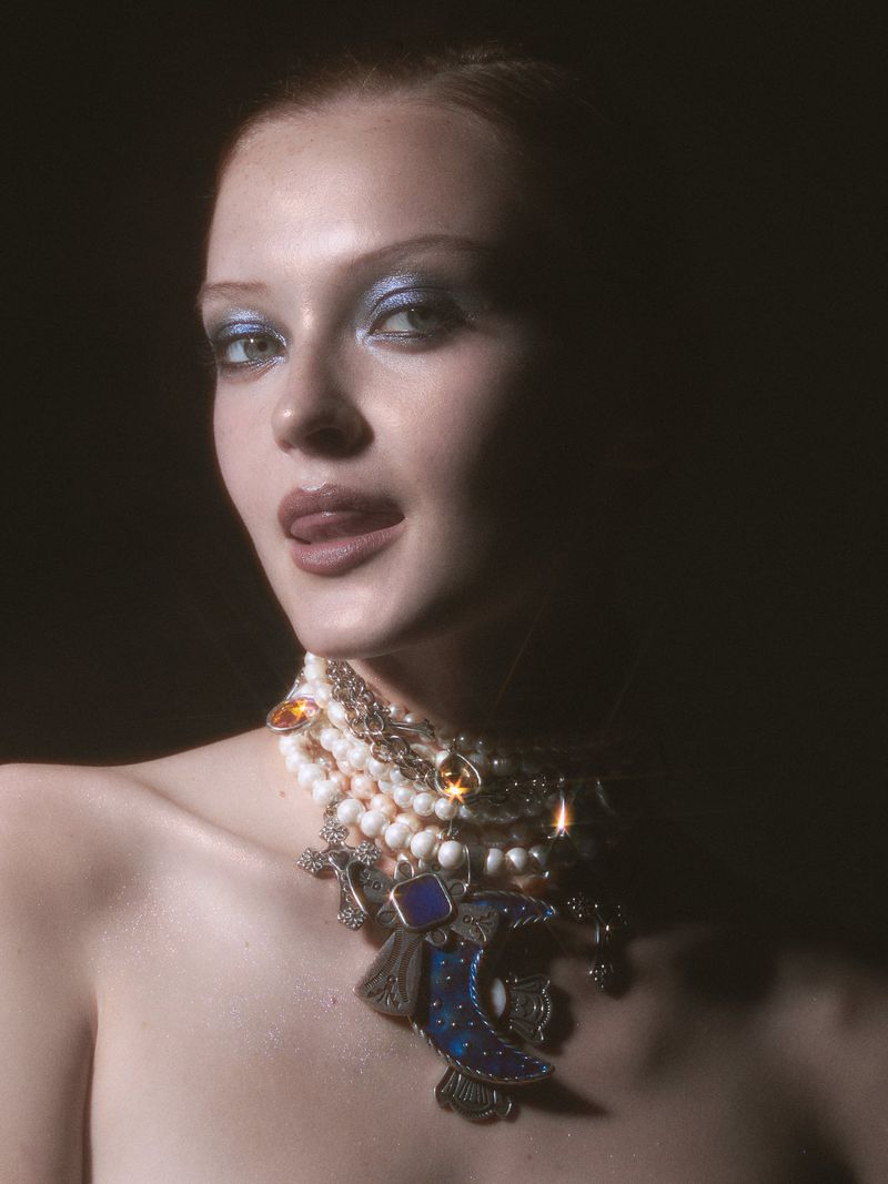 Close-up portrait of a woman wearing a heavy layered pearl necklace with jeweled pendants and blue eyeshadow.