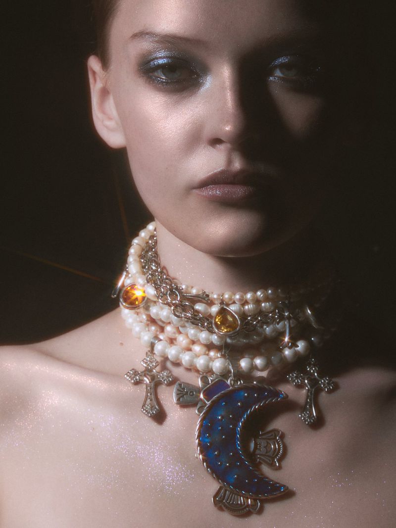 Close-up portrait of a woman wearing a heavy layered pearl necklace with silver cross charms and a large blue crescent moon pendant.