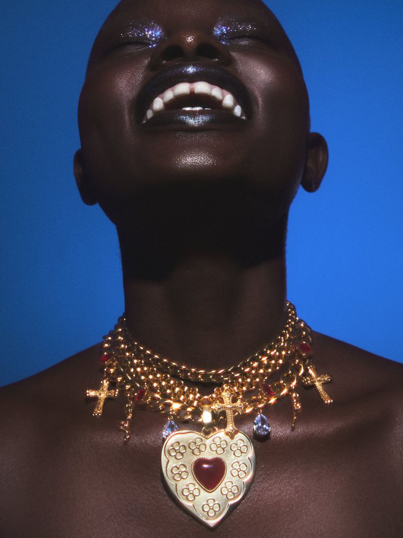 Close-up of a smiling Black woman with glittery eyeshadow and multiple gold cross necklaces against a solid blue background.