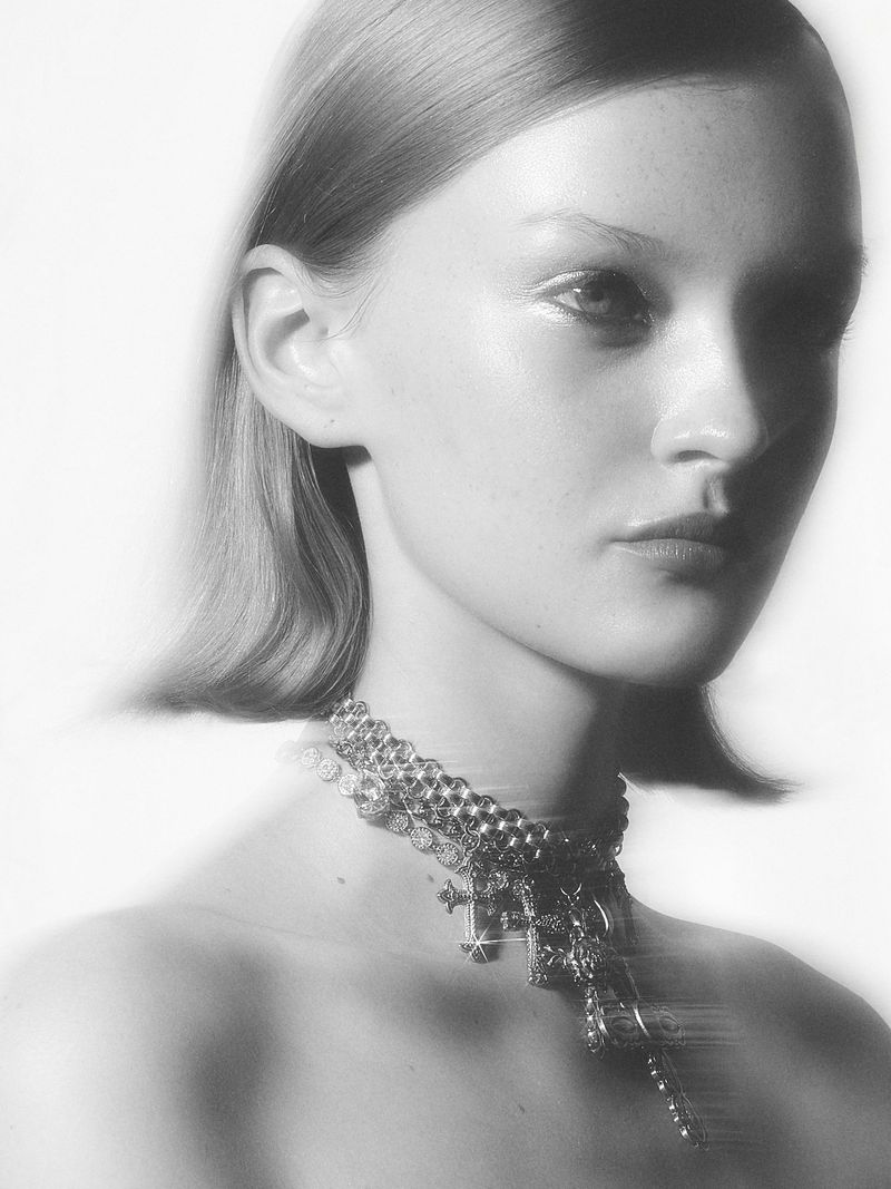 Monochromatic portrait of an adult woman with blonde hair, wearing a layered metal necklace against a blurred light background.