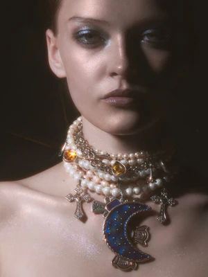 Close-up portrait of a woman wearing a heavy layered pearl necklace with silver cross charms and a large blue crescent moon pendant.