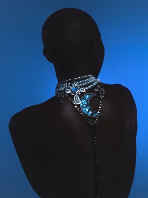 Rear view of a person wearing a pearl choker and ornate crystal cross and crescent moon necklaces against a blue background