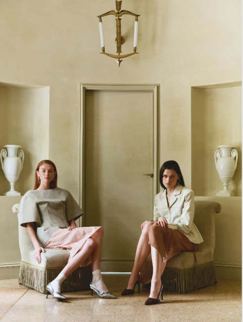 Two young women sit in fringed chairs indoors. The interior is decorated in neutral tones with a chandelier and classic vases.