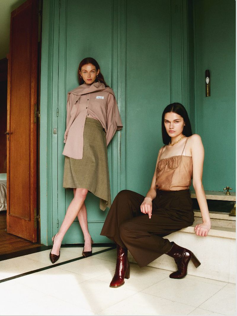 Two fashion models pose indoors against a teal-painted wall. One sits on a marble ledge, the other stands wearing high heels.