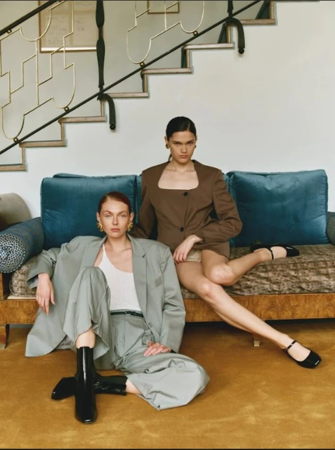 Two young adult women in blazers and skirts pose indoors on a sofa and carpet with neutral wall and stair background. Fashion editorial shot.