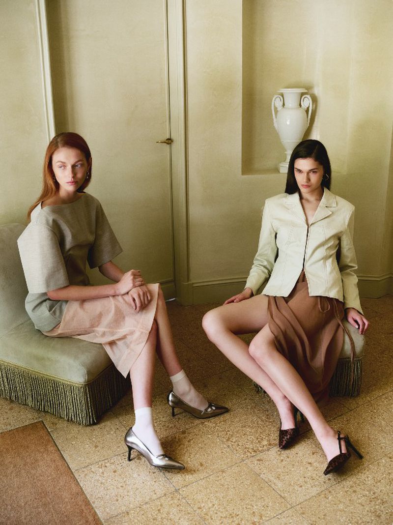 Two fashion models pose indoors, wearing modern clothing. One model is seated on a chaise, the other directly on the floor.