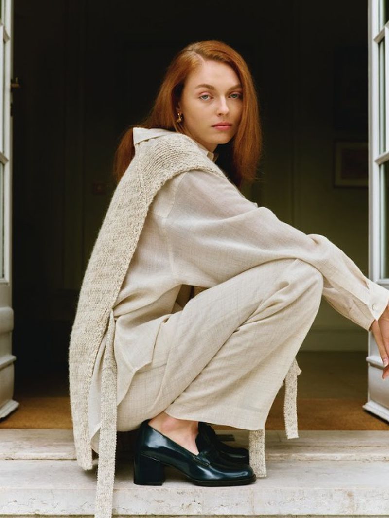 Red-haired woman in relaxed pose wearing casual linen clothing and block-heeled patent-leather loafers squats in front of a doorway.