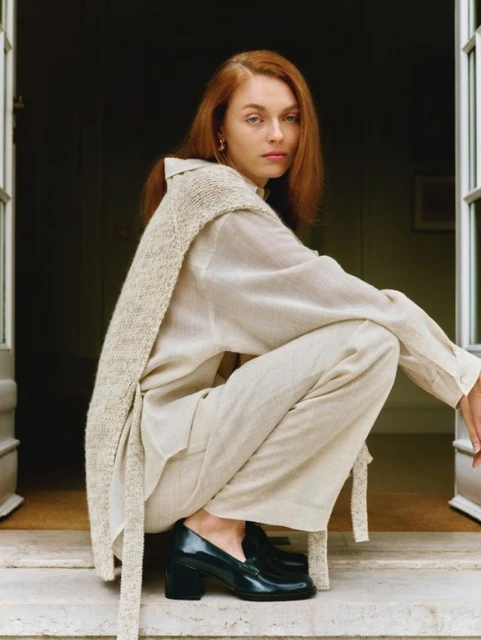 Red-haired woman in relaxed pose wearing casual linen clothing and block-heeled patent-leather loafers squats in front of a doorway.