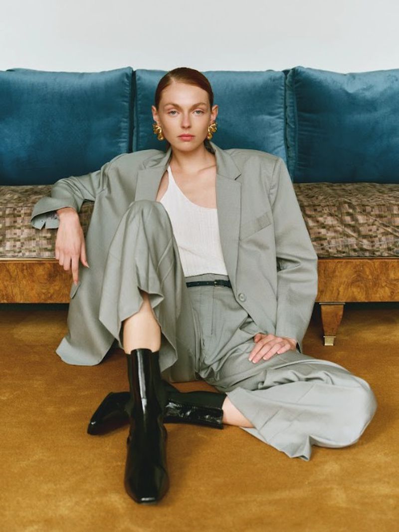 Model wearing an oversized grey suit jacket, matching trousers, white top, and black boots sitting on a golden carpet