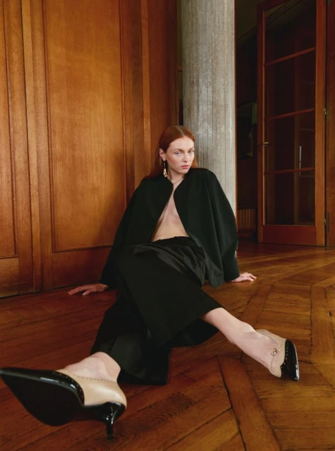 Fashion shot of a red-haired woman in a black outfit and two-tone heels, sitting on a wood floor against a wood wall and grey column.