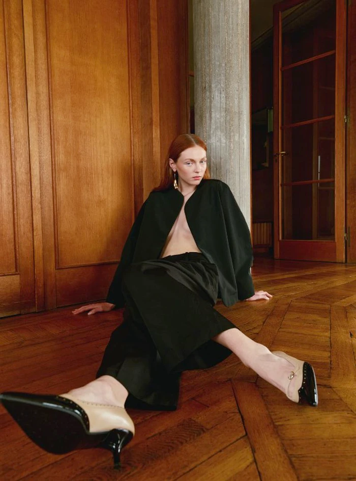 Model with red hair wearing black outfit and pointed-toe kitten heels seated on a wooden parquet floor in a panelled interior.