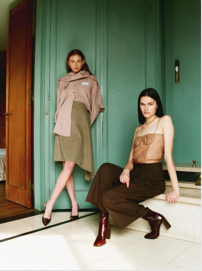 Two female models posing indoors against a green wall, one standing and one seated wearing autumn fashion apparel.