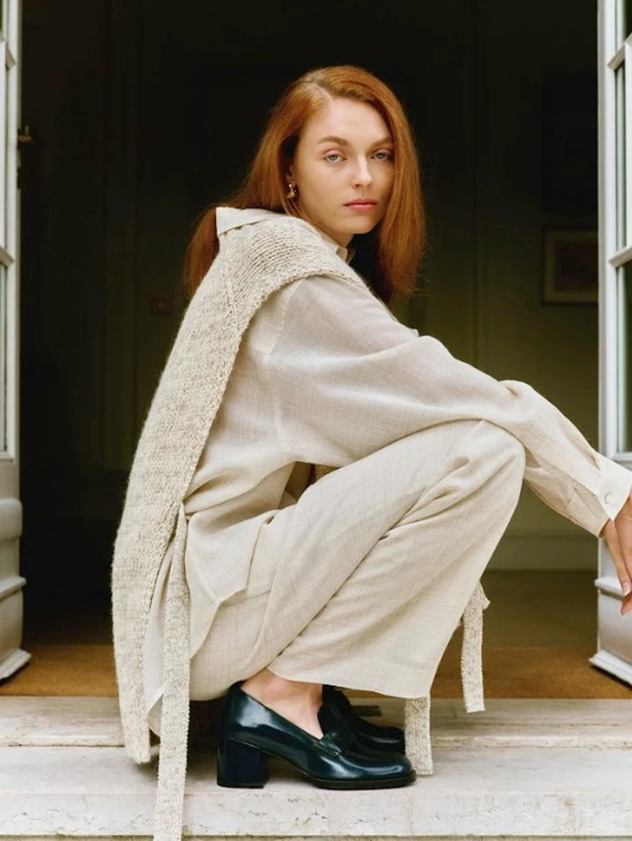 Red-haired model crouching in a doorway wearing a beige shirt, matching trousers, a knitted sweater draped over shoulders, and dark loafers.