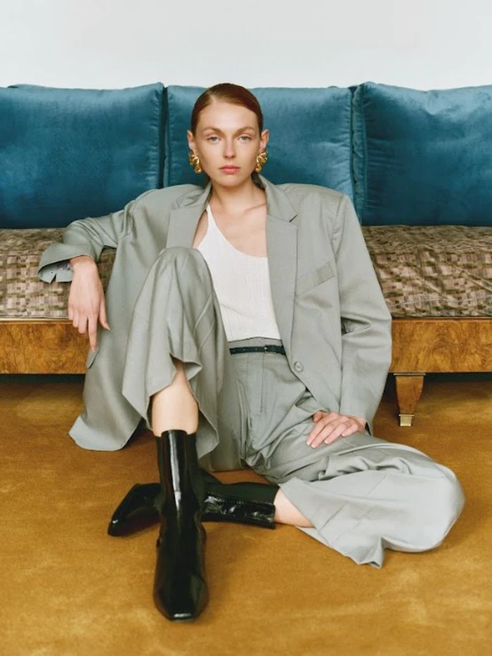 Model wearing an oversized grey suit jacket, matching trousers, white top, and black boots sitting on a golden carpet