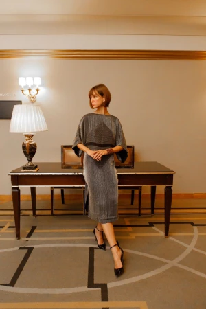 Elegant woman in a metallic dress and heels poses indoors near a dark wood table with decorative lamp, creating a stylish, sophisticated look.