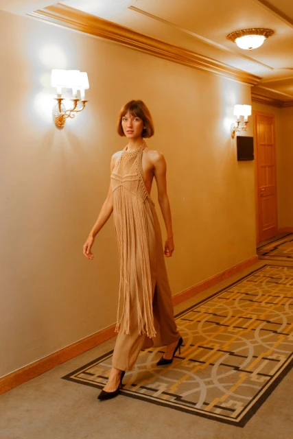 A woman walks in a hallway wearing a sleeveless tan fringe dress with matching pants and heels, walls are tan with patterned carpet.