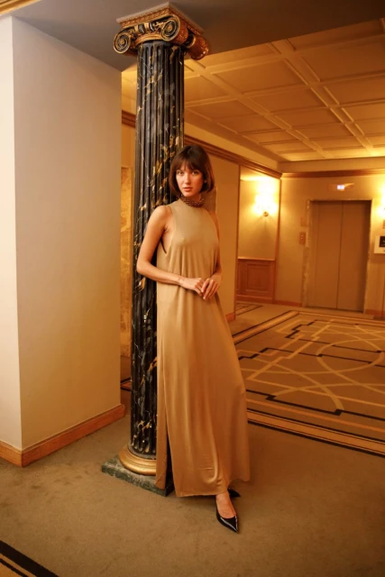 Brunette woman in a long tan dress poses indoors next to a decorative column in a hallway with carpet and an elevator in the background.